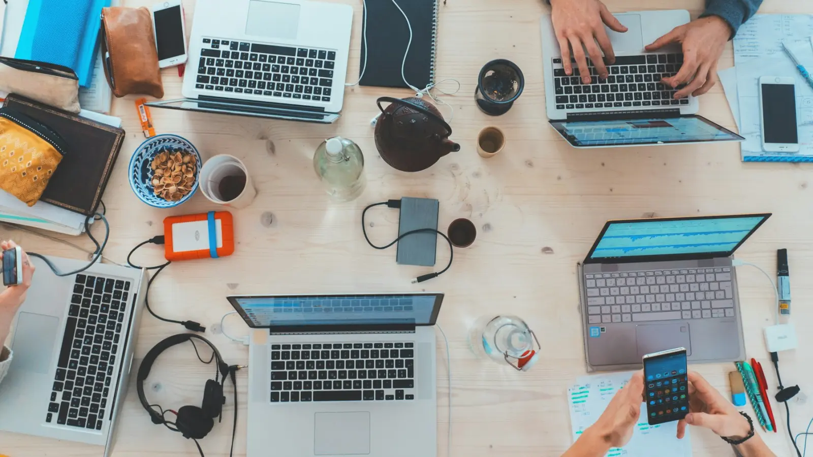 Table de travail vue du dessus : plusieurs ordinateurs portables, téléphones et carnets, mains au clavier