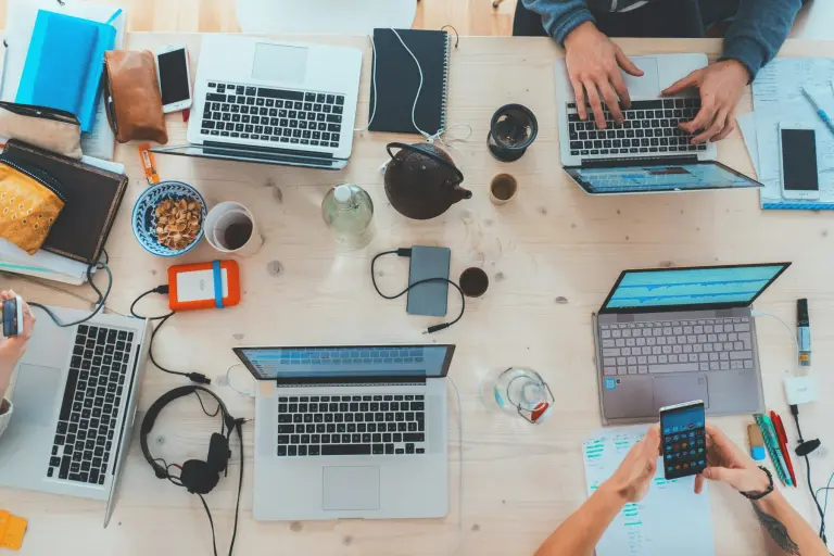 Table de travail vue du dessus : plusieurs ordinateurs portables, téléphones et carnets, mains au clavier